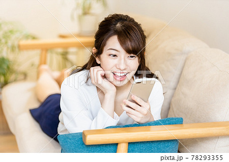 リビングのソファに寝そべってスマホの画面を見る女性 78293355