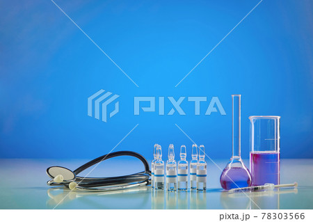 Measuring beaker, medical flask with purple chemical reagent. Six ampoules with liquid, syringe and phonendoscope, blue background. Pandemic COVID-19 78303566