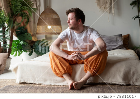 Smiling young man sitting on his bed while looking at the camera Smiling young man sitting on his bed while looking at the camera 78311017