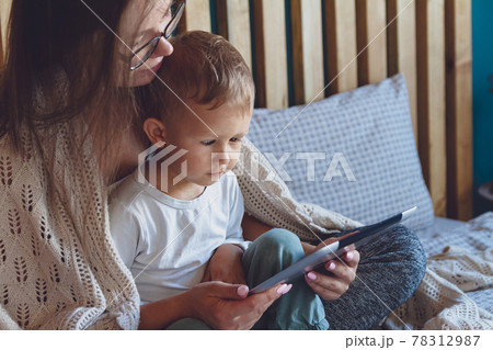 Young mom and son with a tablet under a cozy blanket in the bedr 78312987