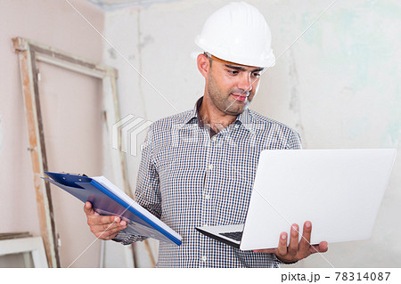Portrait of positive architect working at the notebook in helmet with documents 78314087