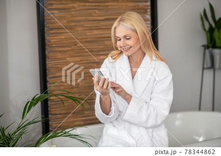 A woman in a white bath robe with a smartphone in her hands 78344862