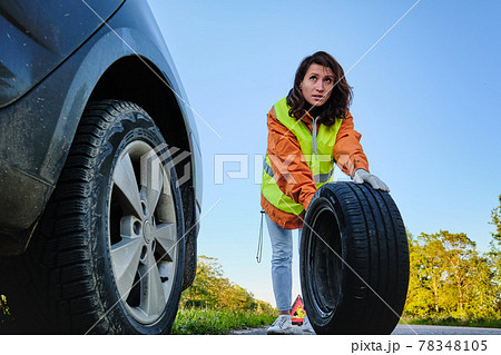The girl changes the damaged wheel of the car. Green vest. Spare wheel. Traffic accident. Travel by car. 78348105