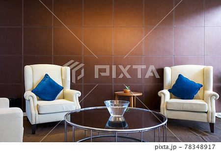Cream armchair and blue cushion in front of a brown leather panel. A glass bowl with water placed on a modern round tempered glass table. 78348917