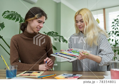 Teenagers guy and girl sitting at table with paints, brushes, drawings Teenagers guy and girl sitting at table with paints, brushes, drawings 78354801