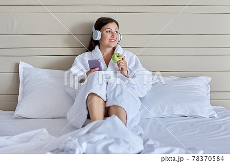 Mature woman relaxing in bed in headphones with smartphone, green apple Mature woman relaxing in bed in headphones with smartphone, green apple 78370584