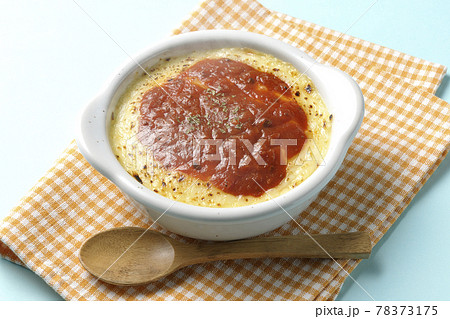 ランチョンマットと耐熱皿に入った溶けて焦げ目がついたたっぷりチーズのドリア、グラタンと木製のスプーン 78373175