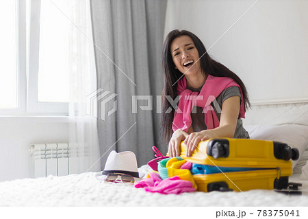 Happy Asian woman trying to closing yellow suitcase full of different clothes accessories and shoes Happy Asian woman trying to closing yellow suitcase full of different clothes accessories and shoes 78375041