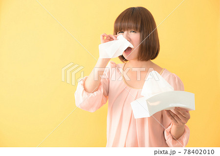 ティッシュボックスを持ちながらつらそうな顔で鼻をかむ若い女性 78402010