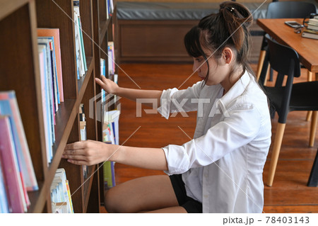 Female college student sitting near bookshelves and looking for a book in the library. 78403143