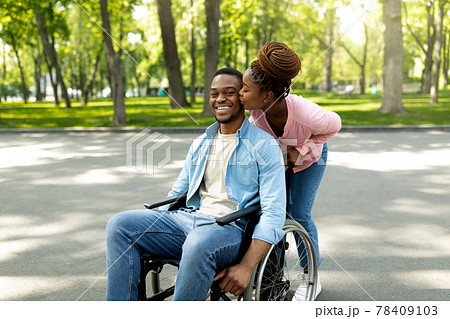 Affectionate black woman kissing her disabled husband in wheelchair, going for walk with him at city park 78409103