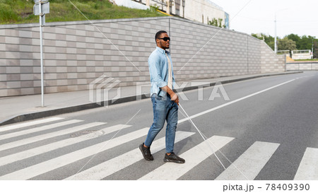 Young black visually impaired man wearing dark glasses, walking across city street, using cane Young black visually impaired man wearing dark glasses, walking across city street, using cane 78409390