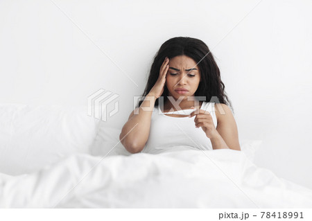 High fever, feeling unwell. Ill black woman holding and looking at thermometer, checking body temperature in bed High fever, feeling unwell. Ill black woman holding and looking at thermometer, checking body temperature in bed 78418991