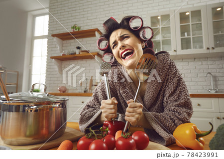 Funny crazy cheerful woman standing in kitchen and holding kitchenware Funny crazy cheerful woman standing in kitchen and holding kitchenware 78423991
