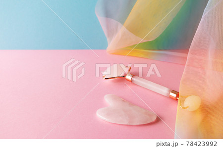 Gua sha and quartz facial roller for skincare massage on blue pink background with colorful rainbow organza drapery 78423992