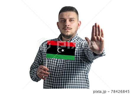 White guy holding a flag of Libya and with a serious face shows a hand stop sign isolated on a white background White guy holding a flag of Libya and with a serious face shows a hand stop sign isolated on a white background 78426845