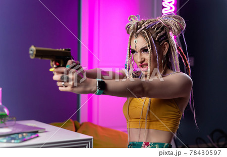 a young hooligan woman with dreadlocks in bright clothes with a gun expresses emotions in neon light 78430597