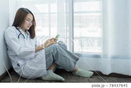 The doctor is wearing a stethoscope Sitting near the window and use the smartphone 78437645