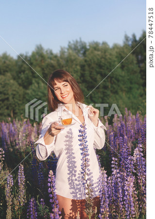 Woman in white shirt in field of lupins and holds glass cup of floral tea in summer Woman in white shirt in field of lupins and holds glass cup of floral tea in summer 78447518