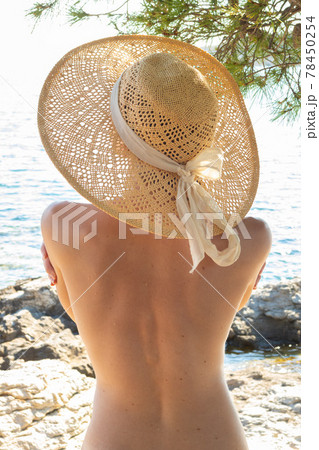 Rear view of topless beautiful woman wearing nothing but straw sun hat realaxing on wild coast of Adriatic sea on a beach in shade of pine tree. FKK sunbathing lifestyle concept. 78450254