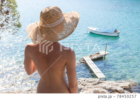 Rear view of topless beautiful woman wearing nothing but straw sun hat realaxing on wild coast of Adriatic sea on a beach in shade of pine tree. FKK sunbathing lifestyle concept. 78450257