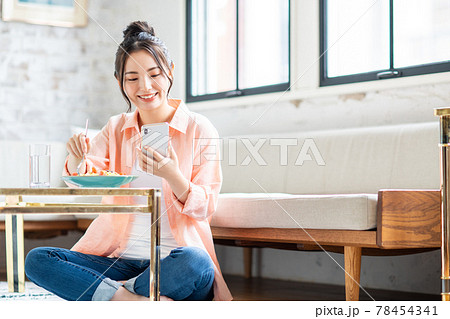 リビングでパスタを食べながらスマホを見る若い女性 78454341
