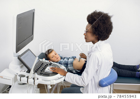 Ultrasound examination of the child in modern clinic. Young afro american female confident doctor makes an abdominal scan of a school 12-aged boy in a medical clinic. Ultrasound examination of the child in modern clinic. Young afro american female confident doctor makes an abdominal scan of a school 12-aged boy in a medical clinic. 78455943
