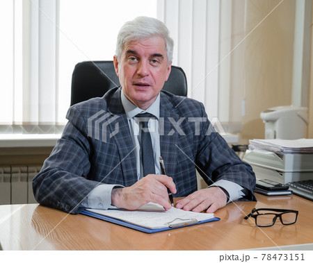 Confident elderly gray-haired man in a plaid suit sits at his desk in the office 78473151
