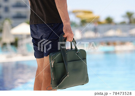 Man tourist in shorts holds business bag in background of hotel pool 78473528