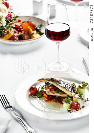 grilled sea bass with sauteed tomatoes, served with a potato ball on a white plate, sea bass fillet plan on a light background, grilled fish fillet served with a glass of wine 78487373