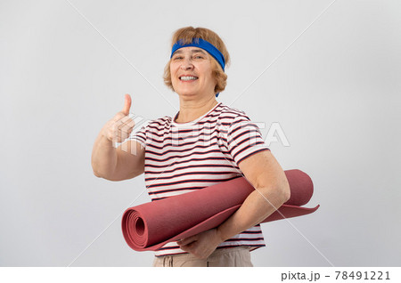 An elderly woman holds a yoga mat and shows her thumb up. The pensioner approves of sports. An elderly woman holds a yoga mat and shows her thumb up. The pensioner approves of sports. 78491221