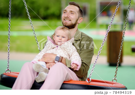 Toddler girl having fun on outdoor playground. Young father rides daughter on swing. Spring/summer/autumn active leisure for family with kids. 78494319