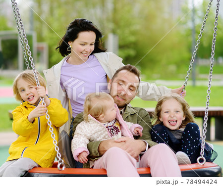 Merry family with three little children having fun on outdoor playground. Young parents rides siblings on swing. Spring/summer/autumn active leisure for family with kids. 78494424