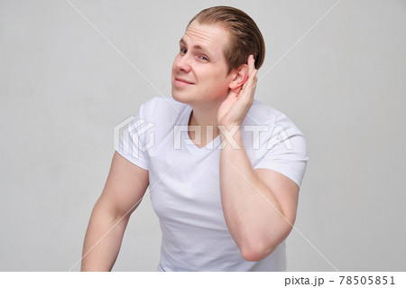 A young man in a white T-shirt can't hear well. The hand is near the ear. A young man in a white T-shirt can't hear well. The hand is near the ear. 78505851