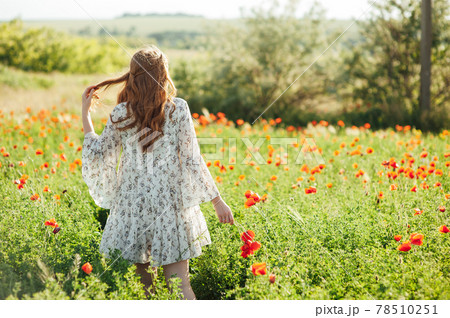 Girl wearing white dress and straw hat walks among red poppy flowers on the sunset. 78510251