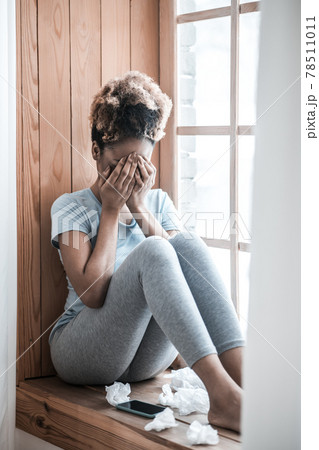Darkskinned woman covering face with hands sitting on windowsill Darkskinned woman covering face with hands sitting on windowsill 78511011