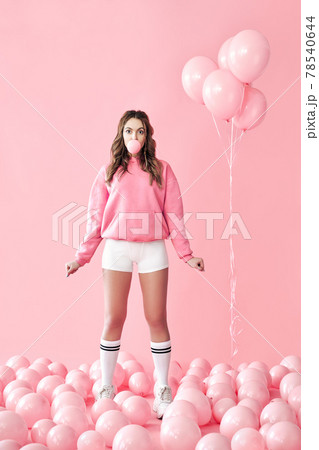Young playful woman blowing bubble with chewing gum staying in pink air balloons background 78540644