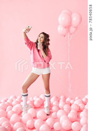 Full length portrait of trendy young woman making selfie photo on smartphone holding milk shake over pink balloons background Full length portrait of trendy young woman making selfie photo on smartphone holding milk shake over pink balloons background 78540658