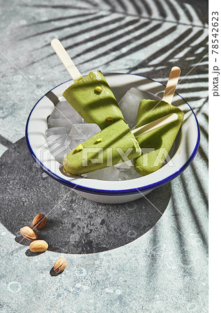 Frozen homemade pistachio popsicle in bowl of ice on gray background with palm leaf shadow in harsh light. Refreshing popsicle, frozen green juice on stick. Top view, copy space Frozen homemade pistachio popsicle in bowl of ice on gray background with palm leaf shadow in harsh light. Refreshing popsicle, frozen green juice on stick. Top view, copy space 78542623
