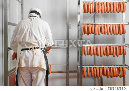 Worker hangs raw sausages on racks in storage room at meat processing factory 78548550