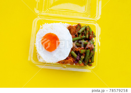 Rice topped with spicy stir fried pork and yard long bean with red curry paste, fried egg in plastic food container on yellow background. Rice topped with spicy stir fried pork and yard long bean with red curry paste, fried egg in plastic food container on yellow background. 78579258