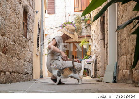 Beautiful blonde young female traveler wearing straw sun hat caressing domestic cat while enjoying summer vacation in an old traditional costal town at Adriatic cost, Croatia. 78593454