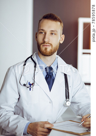 Friendly bearded doctor sitting and writing at clipboard in sunny clinic. Medicine concept 78595878