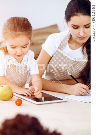 Happy woman and her daughter are searching for new recipes for a family feasting, sunny kitchen. Christmas, New year, Thanksgiving, Anniversary, Mothers Day. Healthy meal cooking concept Happy woman and her daughter are searching for new recipes for a family feasting, sunny kitchen. Christmas, New year, Thanksgiving, Anniversary, Mothers Day. Healthy meal cooking concept 78612963