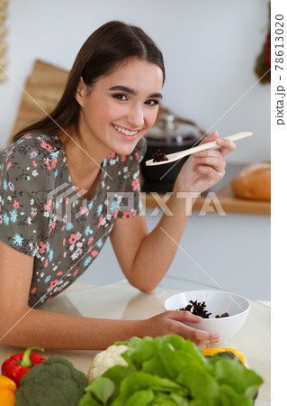 An attractive young dark-haired woman tastes a new recipe for a delicious salad mix while sitting at the table in the kitchen 78613020