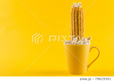 A yellow ear of corn sticking out of a yellow cup, as if growing, popcorn is scattered, on a yellow background, concept A yellow ear of corn sticking out of a yellow cup, as if growing, popcorn is scattered, on a yellow background, concept 78629361