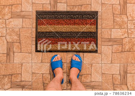 Male feet in blue house slippers stand in front of a foot mat on a brown tiled floor texture background, top view 78636234