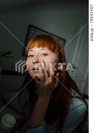 Woman wearing domestic clothes looking at her reflection and applying moisturizer 78645407