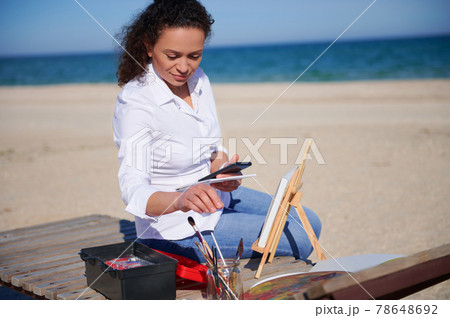 Beautiful woman sitting on a chaise lounge on the beach with smartphone in her hand. Painting tools on the foreground. Beautiful woman sitting on a chaise lounge on the beach with smartphone in her hand. Painting tools on the foreground. 78648692