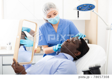 Woman beautician in mask examining face skin of afro american male patient 78657308